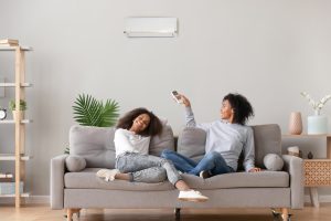 Happy family relax on sofa under air conditioner, mom holding remote control switch on conditioning in living room adjust comfort temperature for daughter, climate system at modern home